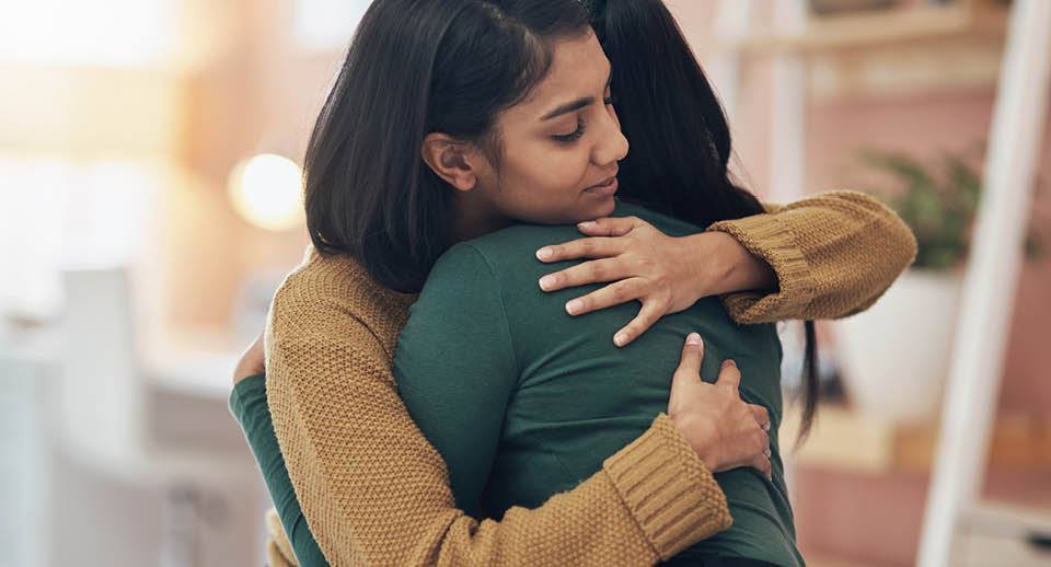 Woman wearing gold sweater comforting woman wearing green shirt.