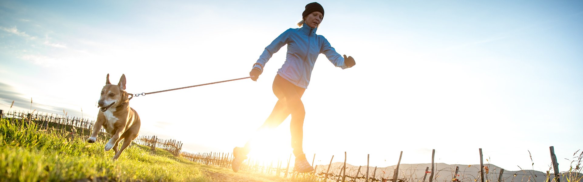 Woman wearing blue jacket walking her dog in the grass at sunrise.